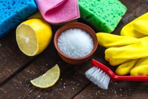 Lemon acid in a small plate, slice of lemon, a juicy lemon, sponge for washing dishes, brushes and yellow rubber gloves for cleaning the house on wooden background